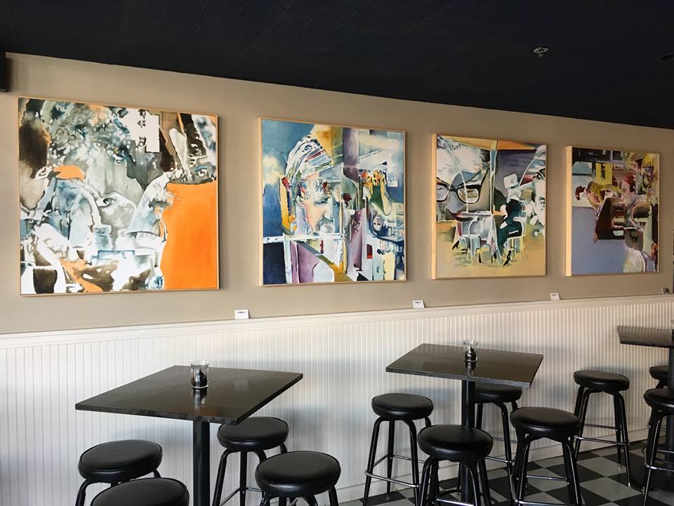 Four 40in square canvas paintings hung on a wall above tables in a restaurant